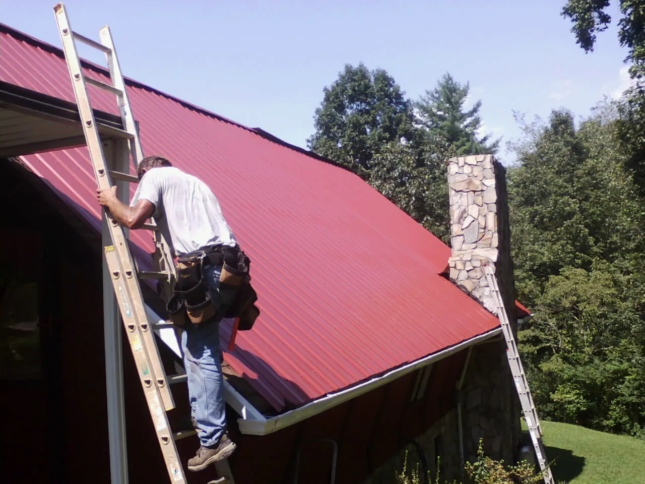 Roofer on a ladder beside a red metal roof and stone chimney — Soffit & Fascia Repair by Roofing Brothers in Schaghticoke