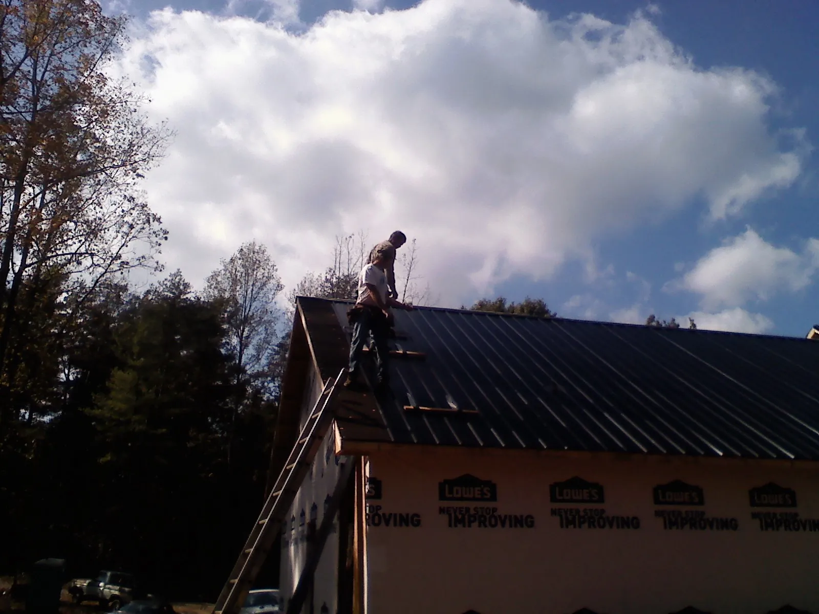 Roofer fastening dark metal panels high against a cloudy sky, from a Roof Inspection job in Schaghticoke